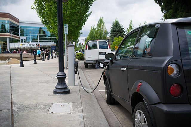 A black car is parked along a sidewalk on a main street, plugged into a charging station. There are trees lining the streets and people walking around in front of business buildings.
