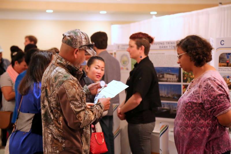People gather at a public information session. There are tables with posters and people engaging. In the foreground, a man examines a paper of information while a woman waits to answer his questions.