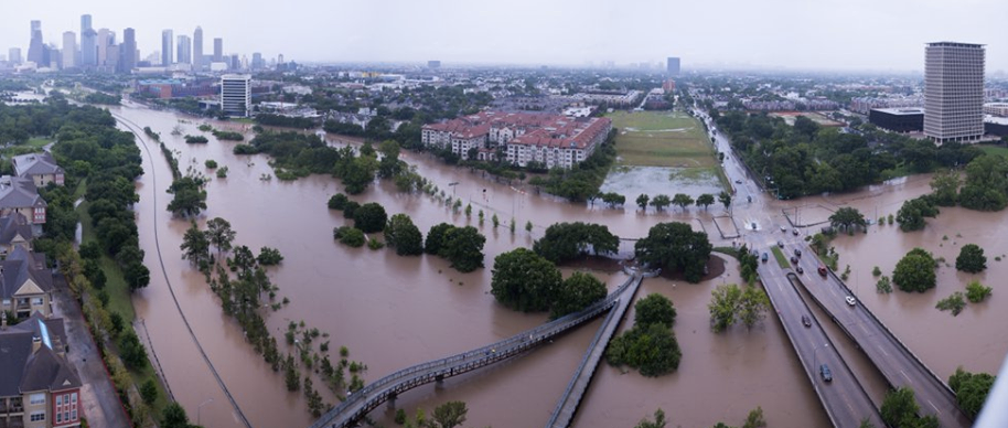 A panoramic birds-eye view of flooding in Houston. Full streets and parks are submerged, and building, trees, and streetlights poke up out of the water.