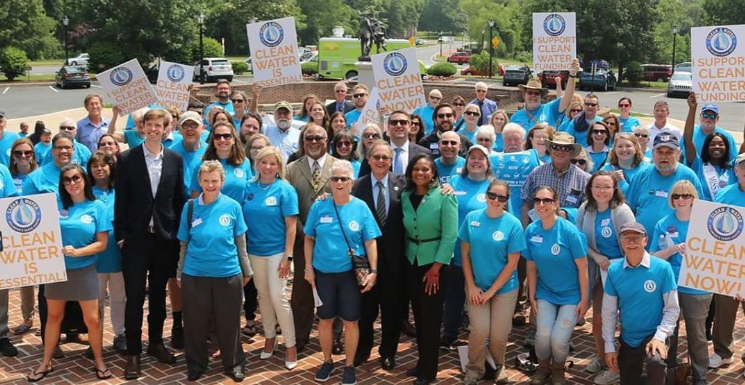 A group of people stand outside wearing matching light blue shirts and smiling up at the camera holding signs that say "Clean Water Now!"