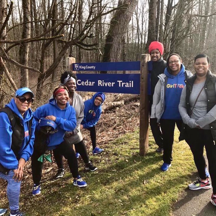 A group of women and girls stand on a trail in the woods, smiling at the camera. They are wearing athletic pants and sweatshirts, ready for a day of hiking. There is a sign that says "King County: Cedar River Trail."