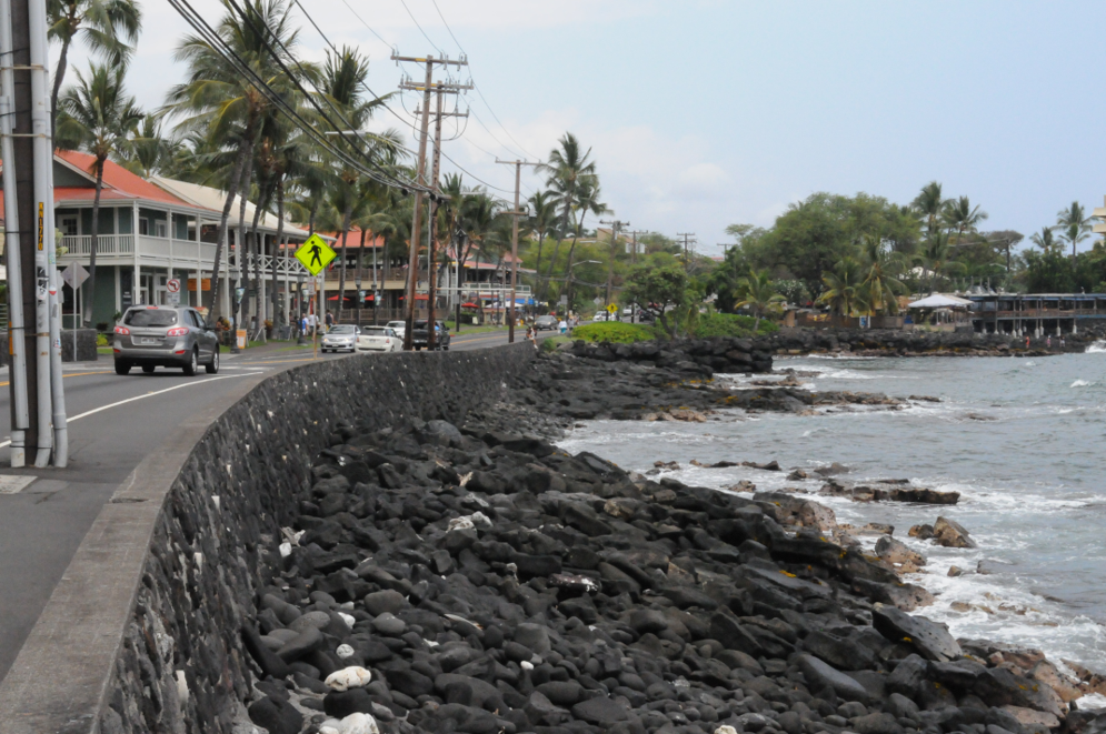 A road runs parallel to a rocky coast, protected only by a short concrete wall.