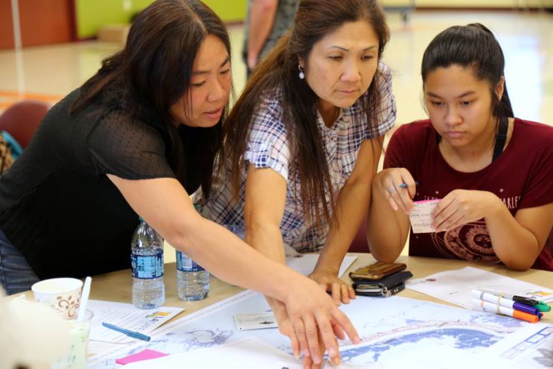Three women sit and stand around a table with a map of a coastal area of Louisiana, talking and gesturing to the map.