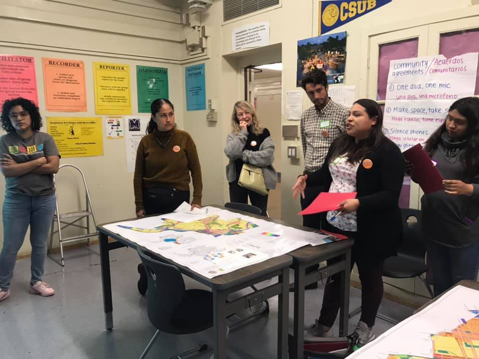 A woman stands at the front of a classroom and speaks animatedly while five other individuals stand beside her and listen. There is a map of the community in front of her and a poster paper behind her that reads "Community agreements" and lists house rules for the meeting. The first three lines read "1. One diva, one mic (una diva, una microfono), 2. Make space, 3. Silence Phones" the rest of the list is not visible.