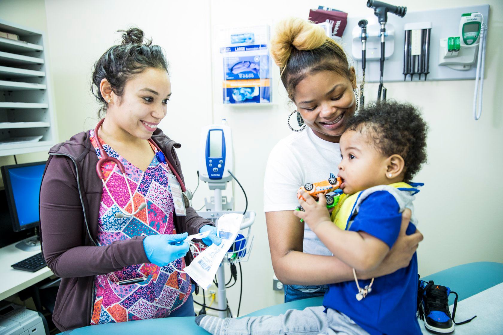 A toddler sits on an examination table held by a smiling woman while a medical professional prepares to perform a checkup.