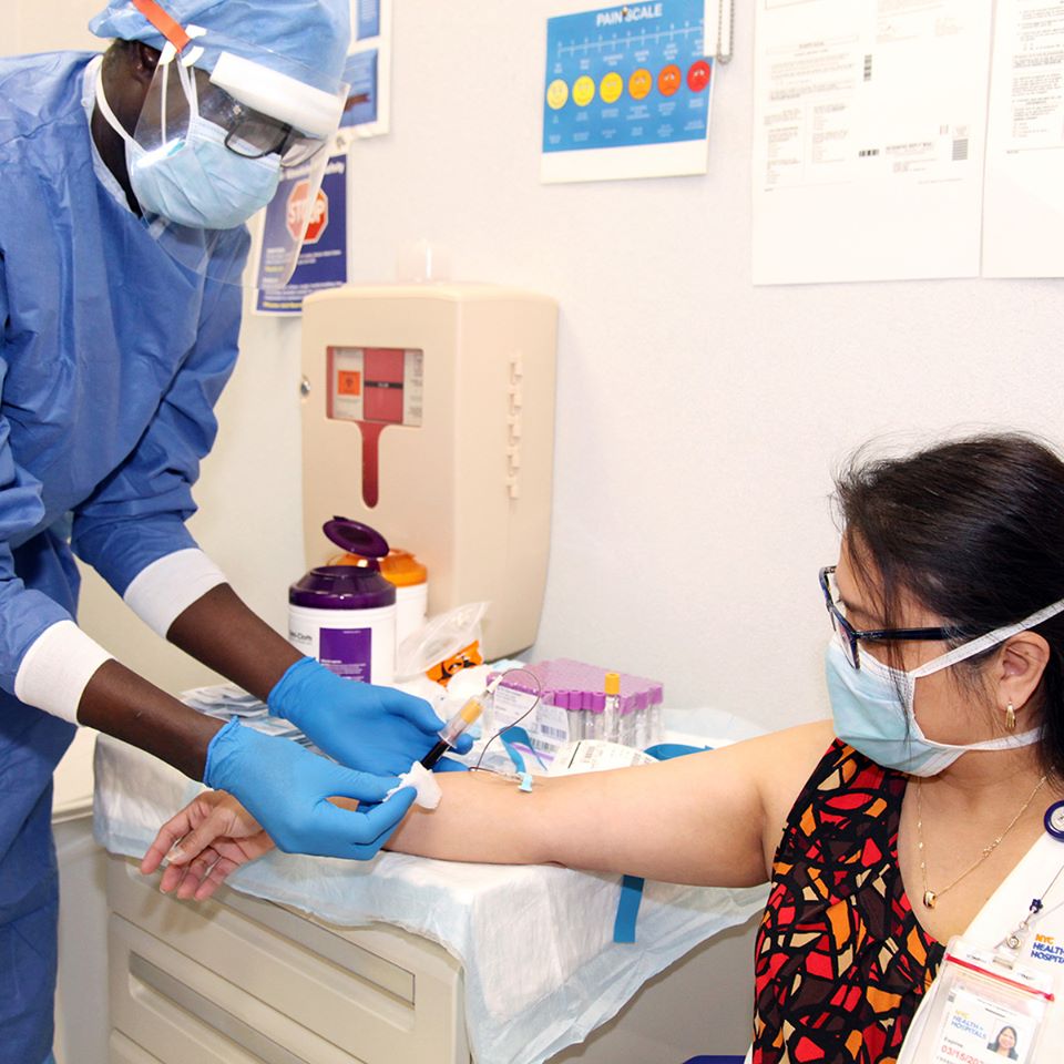A medical professional wears a medical mask, face shield, and protective scrubs while taking a blood sample from a young woman with her arm extended, seated, also wearing a medical mask.