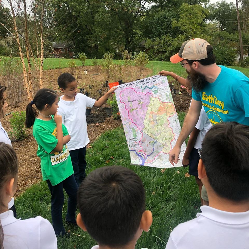 School-age children and an adult educator stand outside near a rain garden and hold up a paper map of the Anacostia Watershed, which they are discussing.