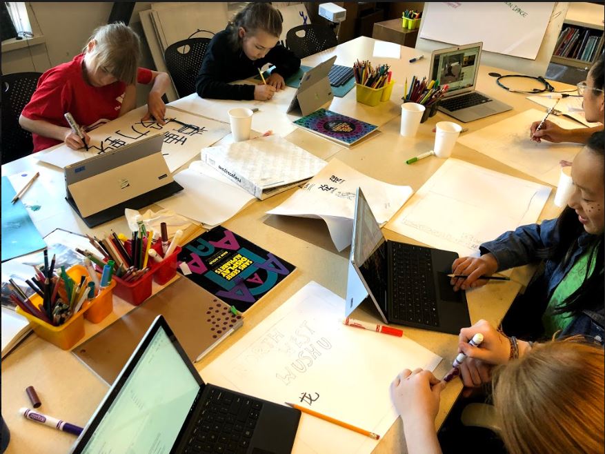Children sit around a table, each with a laptop as well as paper and markers in front of them and work on schoolwork.