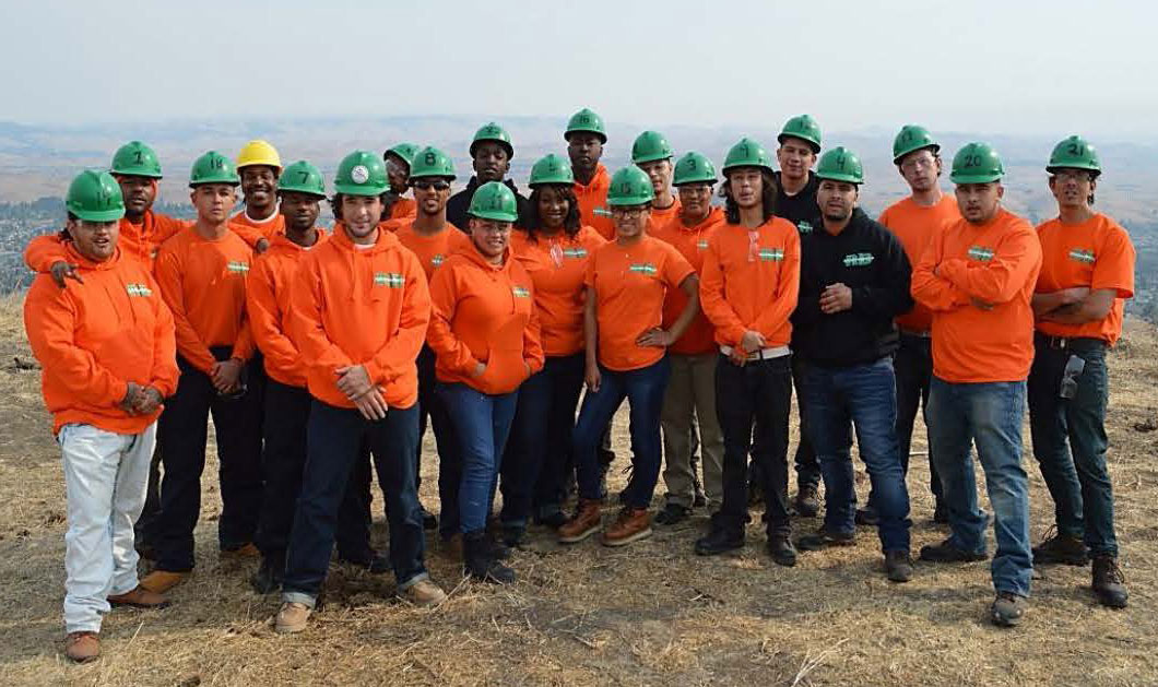 A group of men and women in bright orange shirts and green hardhats stand outside on a hilltop smiling at the camera.