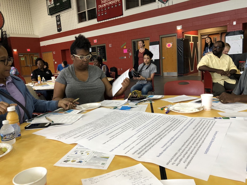 Two women sit at a table as a public listening session in a school gymnasium. They are reviewing a large poster paper titled "Summary of EAG Recommendations"