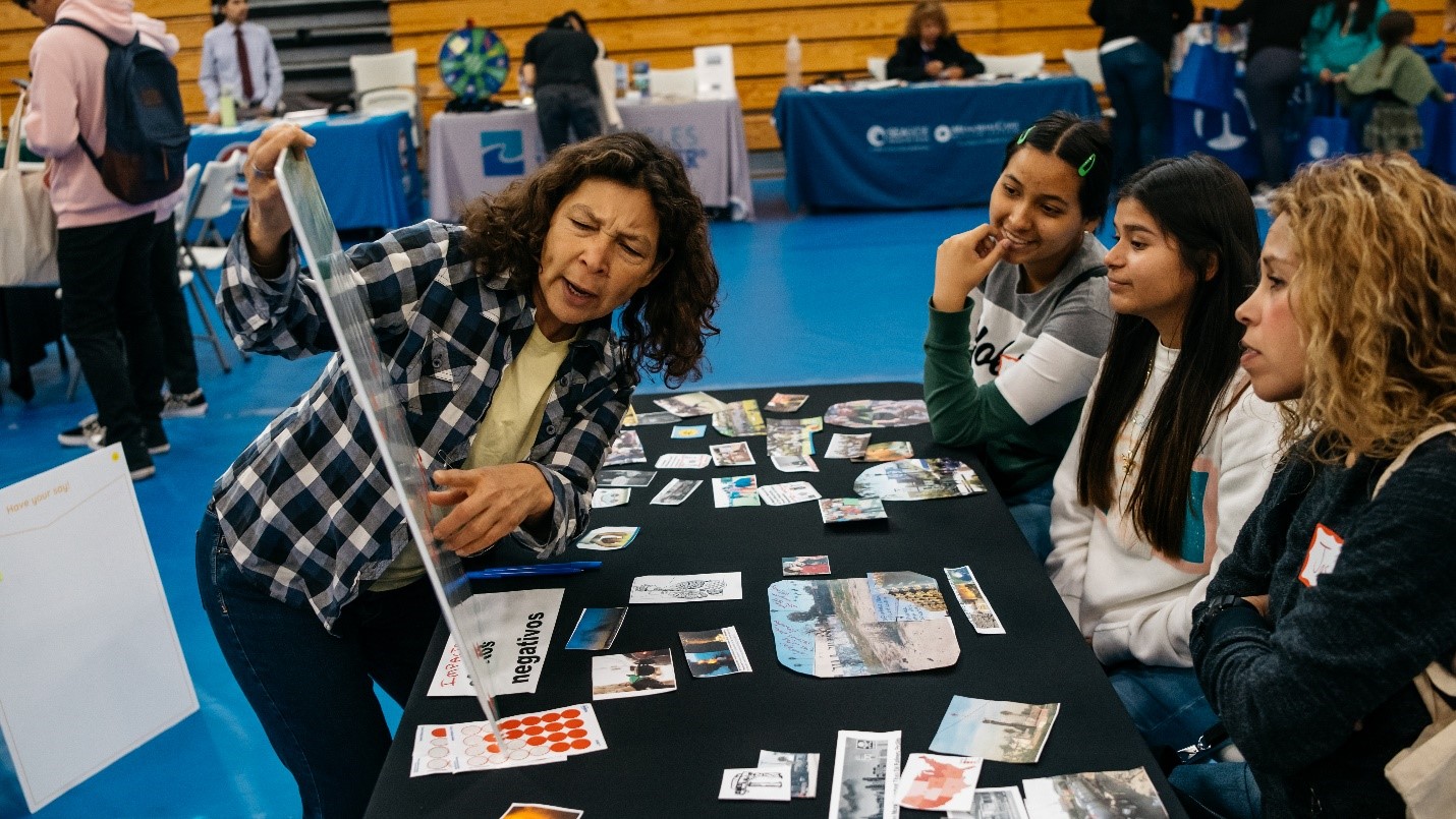 Three women sit on one side of a table with a blue table cloth and an array of pictures on the table. A woman stands on the other side of the table pointing to an informational poster and talking as the other women listen to her.