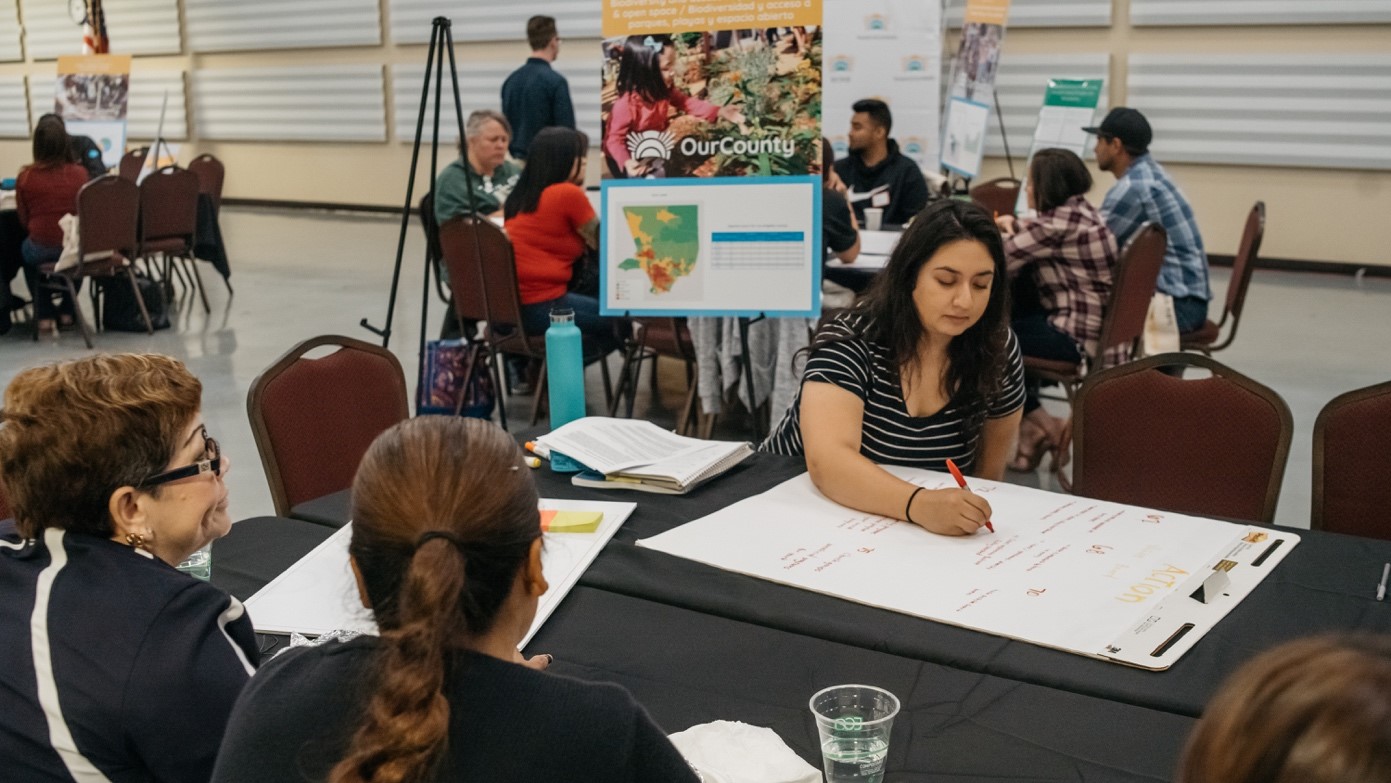 At a public engagement session, four women sit around a table discussing while one writes their ideas down on a poster-sized notepad with a red marker. Behind the woman writing is a poster on a tripod that says, "Our County" and has a picture of a child examining a plant, and a map of the county. There are other tables in the background having similar conversations.