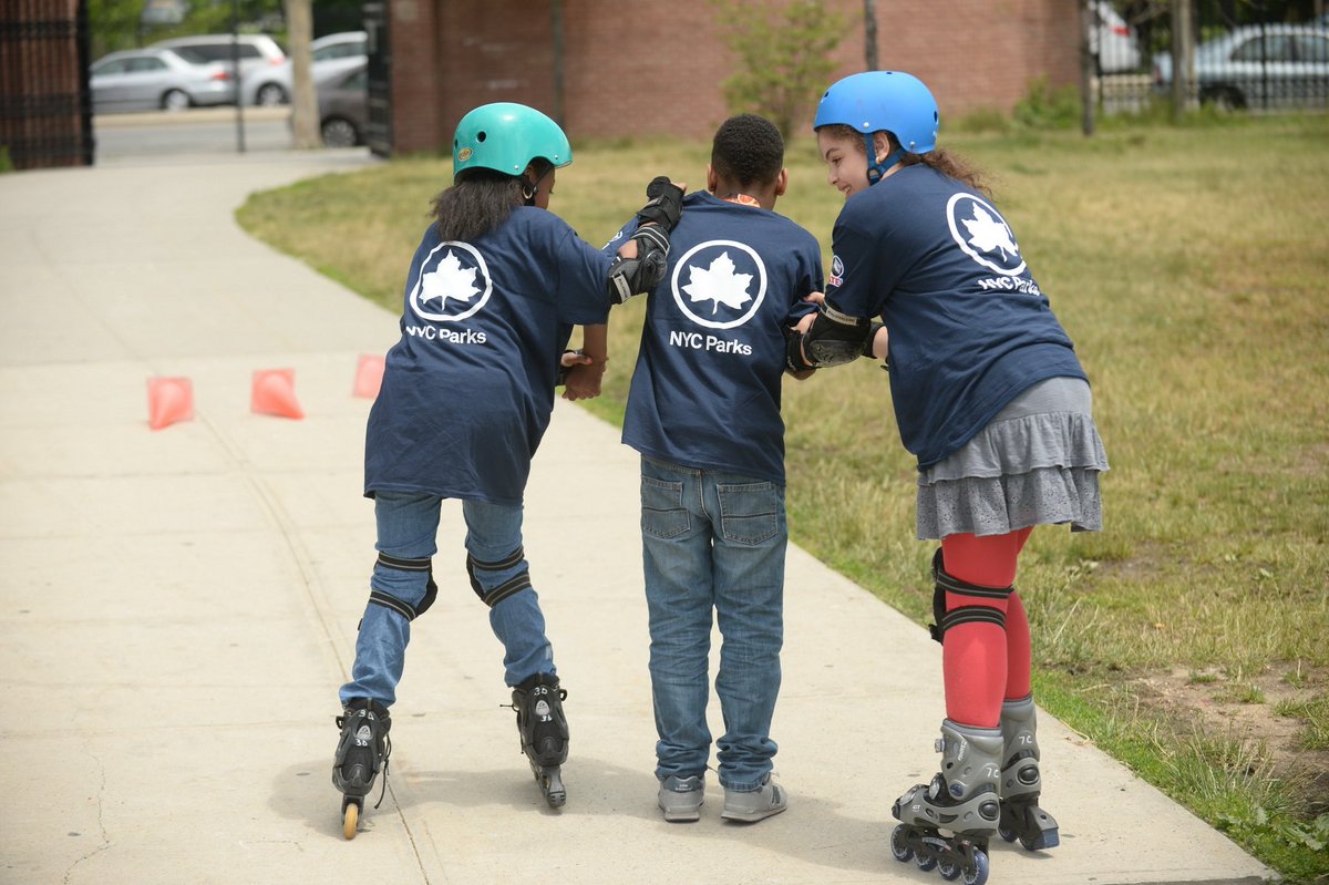 Three children wearing navy blue shirts with white writing that say "NYC Parks" with their backs to the camera. There are two girls on rollerblades and helmets on either side of a boy wearing sneakers.