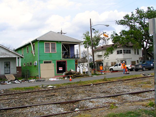 A green house and a white house with broken or empty windows and debris around them. Two people in hard hats and construction vests examine the area.