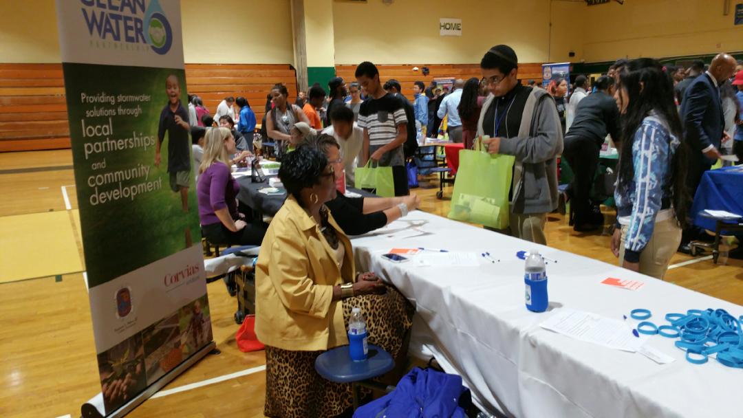 Two women sit behind a table with a white table cloth at a college fair in a school gymnasium. They have a banner behind them that says, "Providing stormwater solutions through local partnerships and community development." They are talking with a high school aged boy and girl who stand on the other side of the table. There are similar conversations going on at other tables in the background.