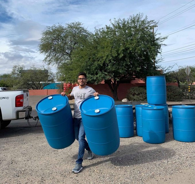 A man carries two empty, large, blue water barrels with stacks of the same type of barrels behind him.