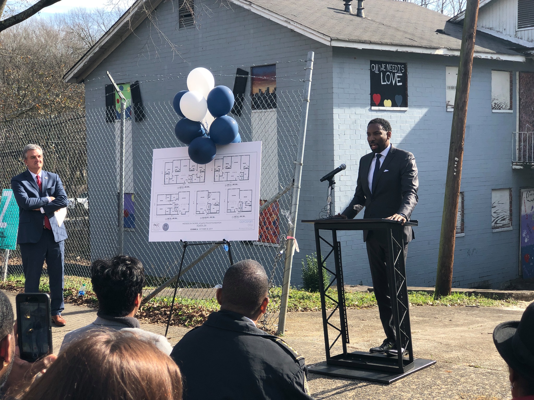 A man stands at a podium outside in front of a group of seated people and addresses the group. There are blue and white balloons tied to a sign next to him.