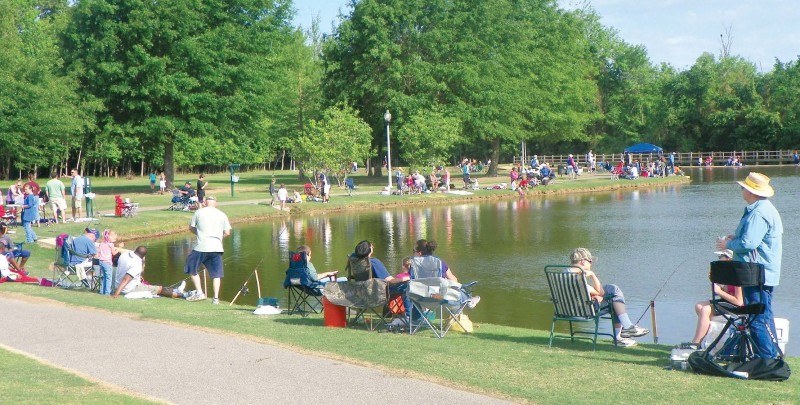 About 50 people in varying couples and family groups enjoy the sunshine on the shore of a lake, some sitting in camp chairs, some fishing.