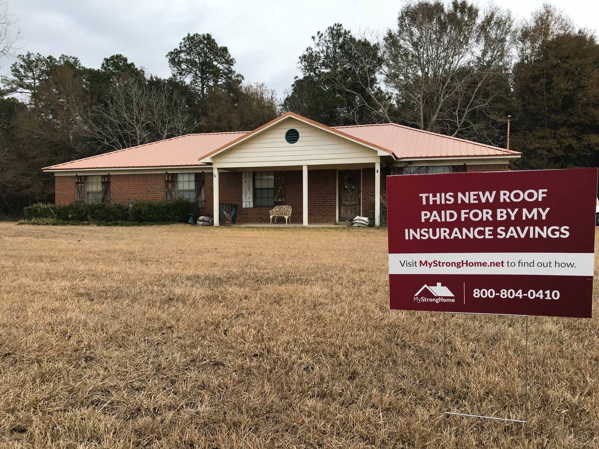 A single-story brick house with a red sign on the front lawn that reads in white lettering: "This new roof paid for by my insurance savings. Visit mystronghome . net to find out how. 800-804-0410."