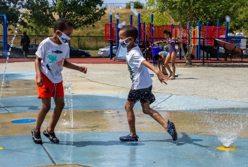 Two young boys play outside in a splash park in Philadelphia. They are wearing shorts, t-shirts, water shoes and cloth masks that cover their noses and mouths. They are running through two small jets of water shooting up from the ground.