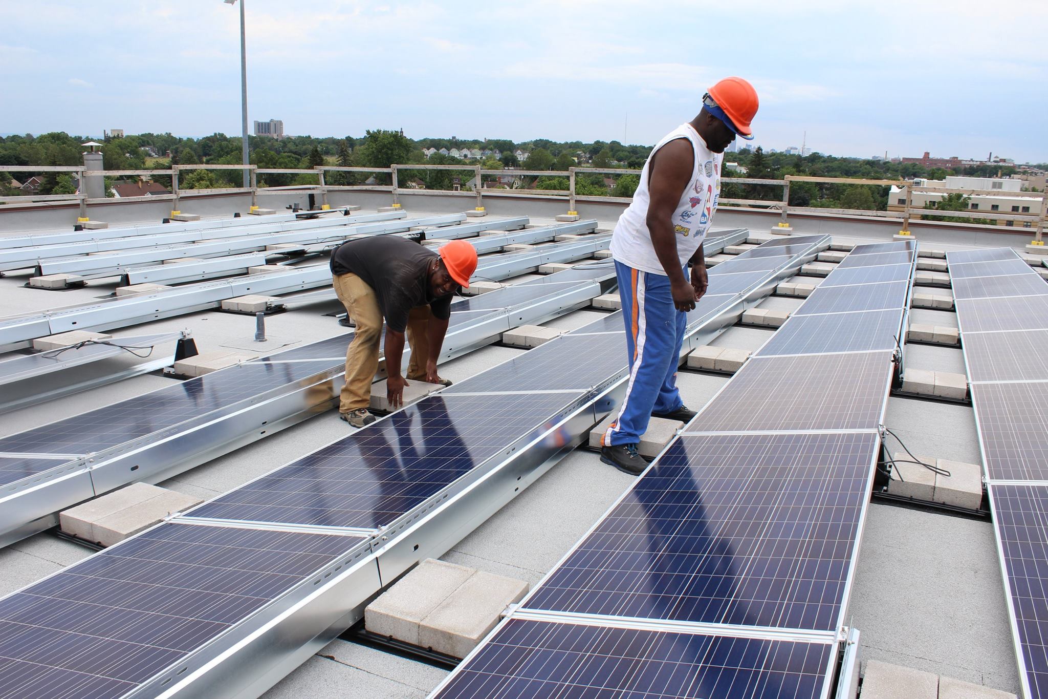Two men working on installing solar panels on a school roof. There are rows of solar panels and the men are bent over working on installing two.