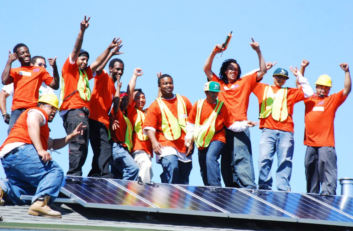 A group of men stand on a rooftop behind a solar panel they just installed. They are wearing orange shirts and construction vests, and raising their arms in a celebratory way.