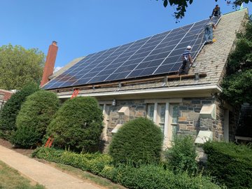 A stone house with a slanted roof and large hedges in front of it. There are solar panels on the roof and three men are attached to cables working on installing them.