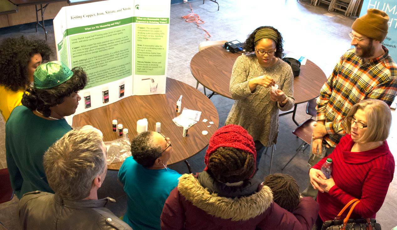 A woman demonstrates how to test water quality samples by holding up a test tube and demonstrating to four people gathered around her.